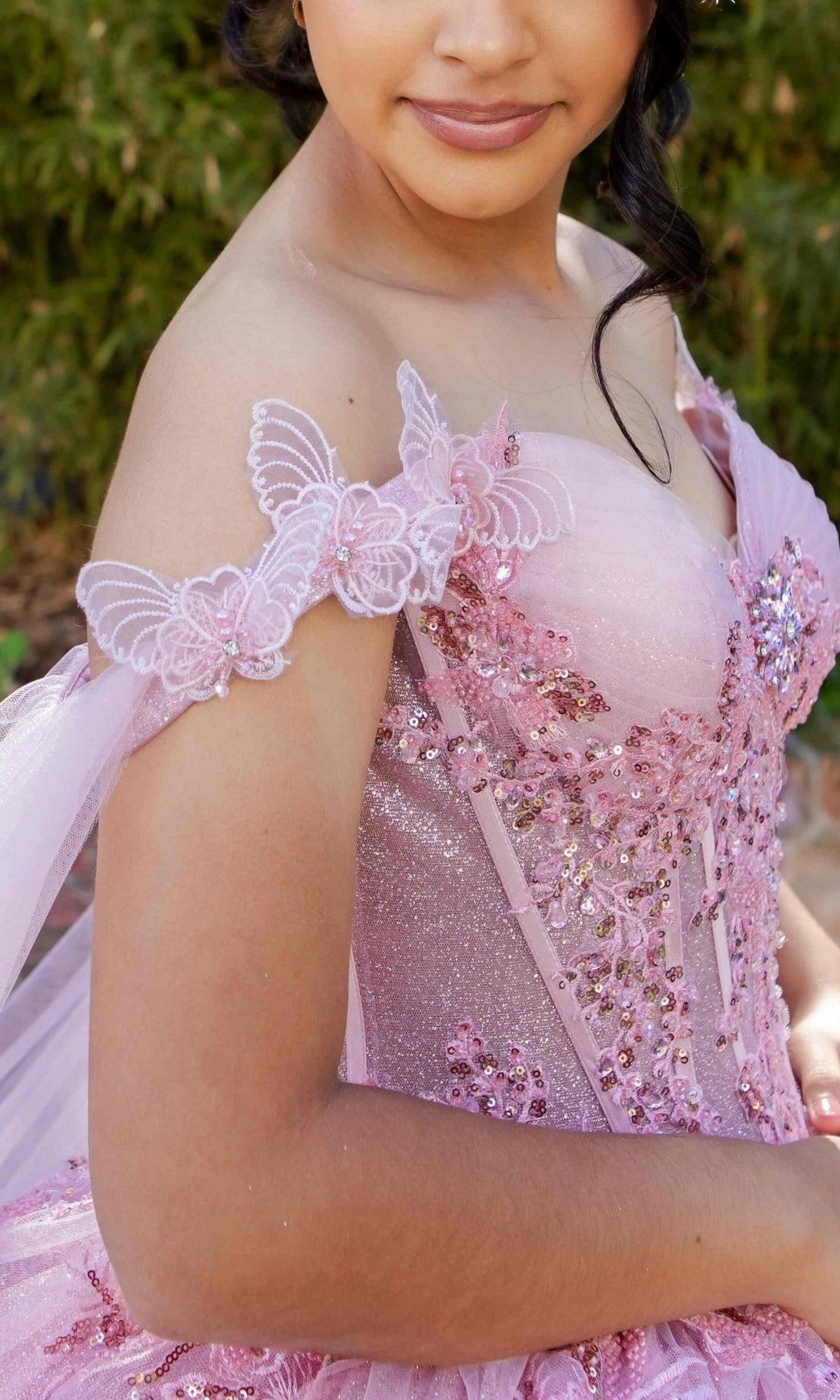 A young person wearing the Quinceanera Dress 8270J By Cinderella Couture—an off-the-shoulder, glittery pink gown adorned with sequins, butterflies, and 3D floral appliques—stands outdoors, showcasing an enchanting ball gown perfect for a magical quinceañera celebration.